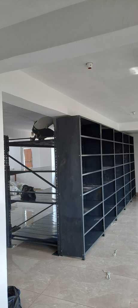 This is a full-height view of the completed shelving unit or the heavy duty pallet rack and the slotted angle rack in Uttarakhand installation. A person can is working on top of the unit, doing installation or adjustment work. The unit is 7 feet tall, reaching nearly to the ceiling, and has multiple sections side by side creating a wall of shelving. The room has modern lighting fixtures including smoke detector on the ceiling. The flooring is finished tile and the walls are painted white. By Rack Manufacturer In Uttarakhand