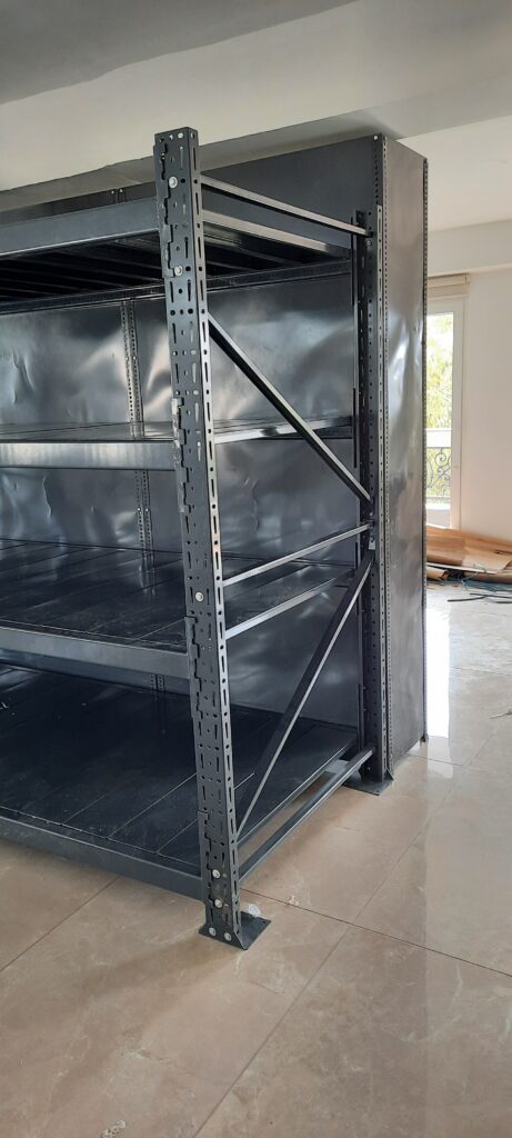 This shows a close-up view of an industrial-style heavy duty pallet rack in Uttarakhand. It's made of dark gray with multiple shelf levels. The upright support post is with many regular holes for adjustable shelf heights. The unit is installed on a tiled floor in a room with white walls. By Rack Manufacturer In Uttarakhand.