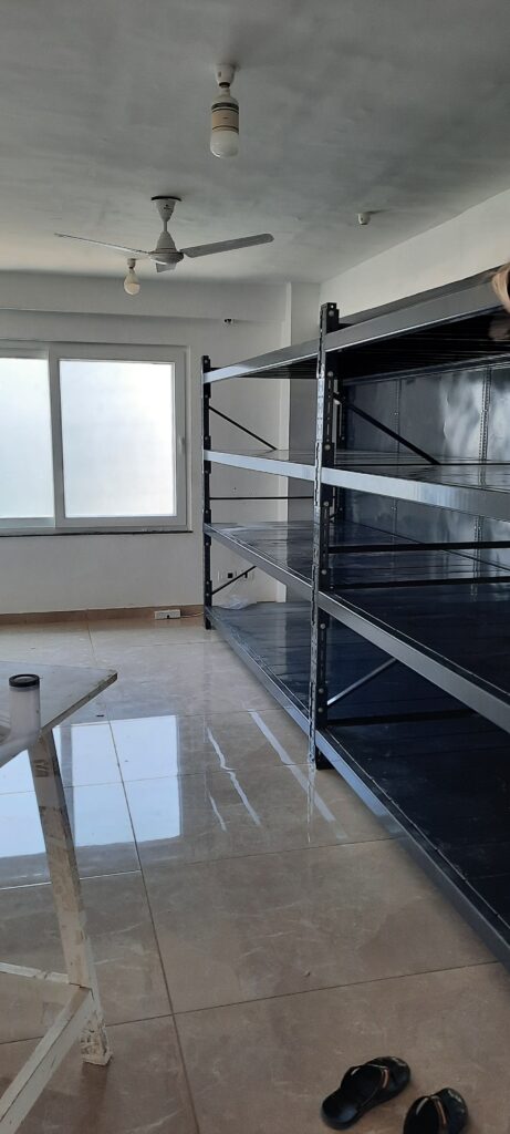 This shows the storage unit from a wider angle. The room has a ceiling fan with a light fixture and additional ceiling lights. There's a window with white frames letting in natural light. The floor is polished tile or marble. The storage unit has multiple levels of shelves with cross-bracing for stability. There appear to be some slippers or sandals on the floor in the foreground.