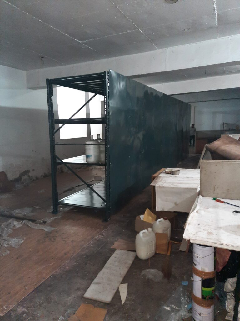 Shelving system, but with some items stored on it, including large water jugs or containers. The shelving unit extends quite long and has side panels installed. The room appears to be under construction or renovation, with some debris on the wooden floor. By Rack Manufacturer In Uttarakhand