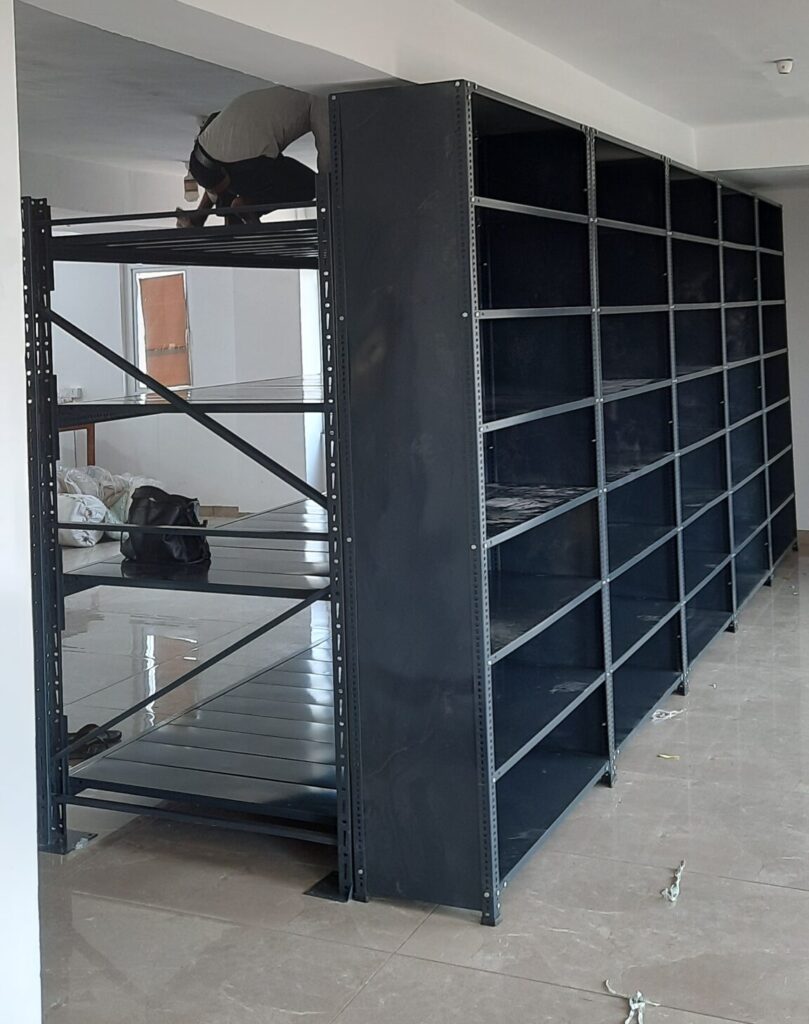 This is a full-height view of the completed shelving unit or the heavy duty pallet rack and the slotted angle rack in Uttarakhand installation. A person can is working on top of the unit, doing installation or adjustment work. The unit is 7 feet tall, reaching nearly to the ceiling, and has multiple sections side by side creating a wall of shelving. The room has modern lighting fixtures including smoke detector on the ceiling. The flooring is finished tile and the walls are painted white. By Rack Manufacturer In Uttarakhand