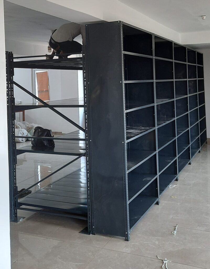 This is a full-height view of the completed shelving unit or the heavy duty pallet rack and the slotted angle rack in Uttarakhand installation. A person can is working on top of the unit, doing installation or adjustment work. The unit is 7 feet tall, reaching nearly to the ceiling, and has multiple sections side by side creating a wall of shelving. The room has modern lighting fixtures including smoke detector on the ceiling. The flooring is finished tile and the walls are painted white.