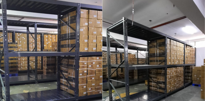 This image depicts a warehouse storage area in the process of installing heavyduty pallet rack. Heavy Duty Pallet Rack with Multi-level steel racking systems, dark gray or black in color, are visible. These racks are partially assembled and extend from floor to ceiling. Inventory Storage on The upper levels of the racks are filled with brown cardboard boxes, neatly stacked and labeled, suggesting organized inventory management. A white ceiling with visible fixtures and a red stripe, likely for utilities or building design. Concrete flooring with visible dust, debris, and materials scattered about. The racking system demonstrates efficient use of vertical space for high-density storage.