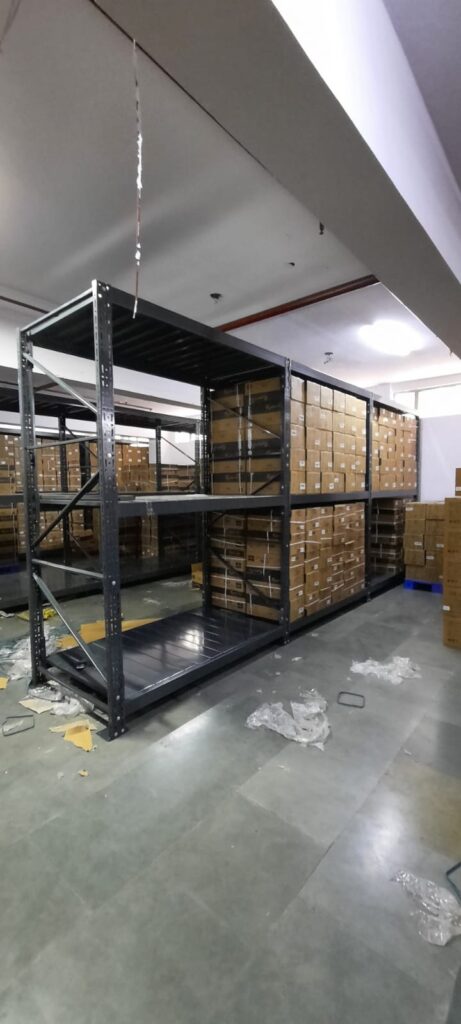 This image depicts a warehouse storage area in the process of installing heavyduty pallet rack. Heavy Duty Pallet Rack with Multi-level steel racking systems, dark gray or black in color, are visible. These racks are partially assembled and extend from floor to ceiling. Inventory Storage on The upper levels of the racks are filled with brown cardboard boxes, neatly stacked and labeled, suggesting organized inventory management. A white ceiling with visible fixtures and a red stripe, likely for utilities or building design. Concrete flooring with visible dust, debris, and materials scattered about. The racking system demonstrates efficient use of vertical space for high-density storage.