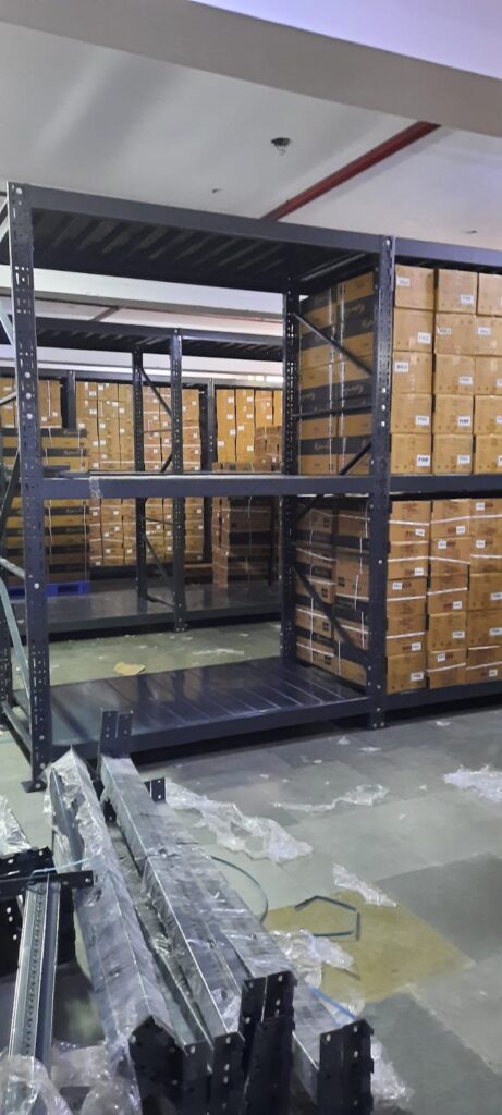 This image depicts a warehouse storage area in the process of installing heavyduty pallet rack. Heavy Duty Pallet Rack with Multi-level steel racking systems, dark gray or black in color, are visible. These racks are partially assembled and extend from floor to ceiling. Inventory Storage on The upper levels of the racks are filled with brown cardboard boxes, neatly stacked and labeled, suggesting organized inventory management. A white ceiling with visible fixtures and a red stripe, likely for utilities or building design. Concrete flooring with visible dust, debris, and materials scattered about. The racking system demonstrates efficient use of vertical space for high-density storage.