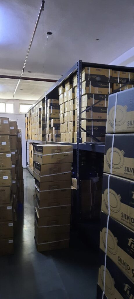 This image depicts a warehouse storage area in the process of installing heavyduty pallet rack. Heavy Duty Pallet Rack with Multi-level steel racking systems, dark gray or black in color, are visible. These racks are partially assembled and extend from floor to ceiling. Inventory Storage on The upper levels of the racks are filled with brown cardboard boxes, neatly stacked and labeled, suggesting organized inventory management. A white ceiling with visible fixtures and a red stripe, likely for utilities or building design. Concrete flooring with visible dust, debris, and materials scattered about. The racking system demonstrates efficient use of vertical space for high-density storage.