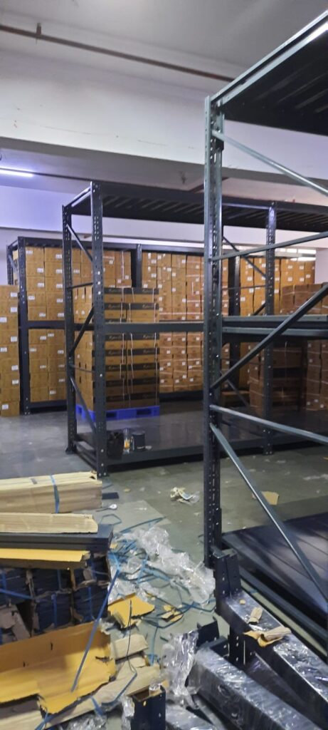 This image depicts a warehouse storage area in the process of installing heavyduty pallet rack. Heavy Duty Pallet Rack with Multi-level steel racking systems, dark gray or black in color, are visible. These racks are partially assembled and extend from floor to ceiling. Inventory Storage on The upper levels of the racks are filled with brown cardboard boxes, neatly stacked and labeled, suggesting organized inventory management. A white ceiling with visible fixtures and a red stripe, likely for utilities or building design. Concrete flooring with visible dust, debris, and materials scattered about. The racking system demonstrates efficient use of vertical space for high-density storage.