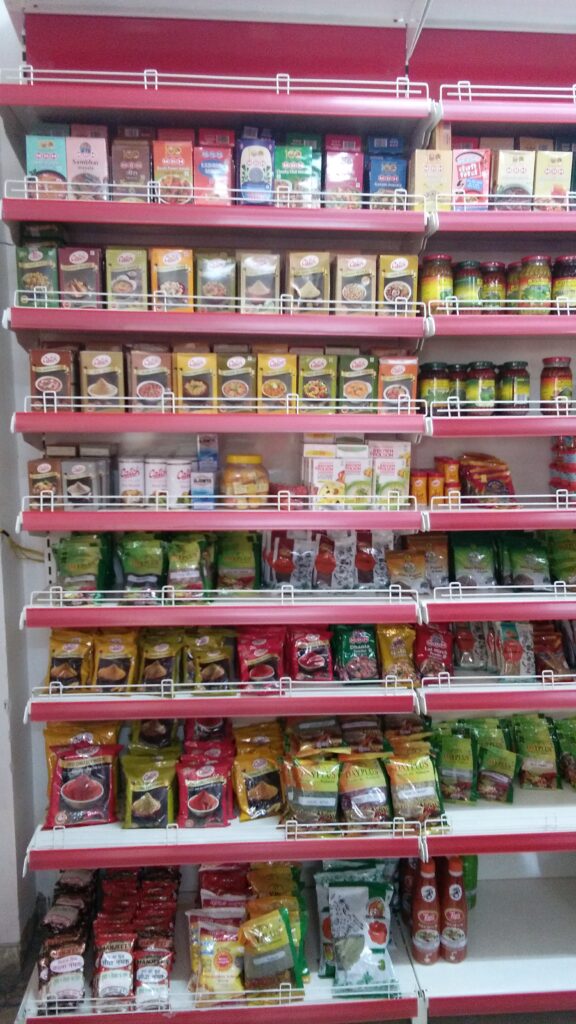 Rack in Himachal Pradesh in a shop showing a closer view of the shelves inside the store. The shelves are stocked with an even wider variety of packaged snacks, canned goods, and other grocery items. The products are organized by category, with similar items grouped together. The shelves are coated in ivory color and with red plastic data strip, creating a visually striking.