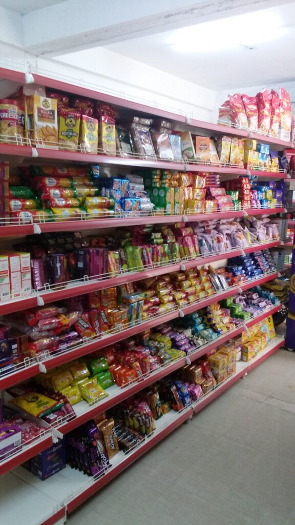 Rack in Himachal Pradesh shop showing a view further down the aisles of the store. The shelves are stocked with a diverse range of products, including cleaning supplies, personal care items, and more packaged snacks and drinks. The store is well-stocked and organized, with a clear effort put into the visual presentation of the merchandise.