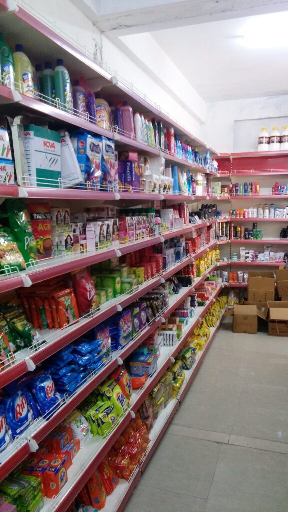 This image shows the interior of a convenience store or small grocery shop. The shelves are stocked with a variety of packaged food and household items. The shelves are coated in ivory color and with red plastic data strip, creating a visually striking. The shelves are organized with various products neatly arranged, suggesting a well-maintained and organized store.