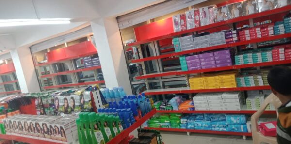 Retail Display Supermarket Rack Supplier And Rack Manufacturer in Andhra Pradesh, This is the interior of a cosmetics or personal care store. The store has bright fluorescent lighting on the ceiling along with ceiling fans. The walls are painted white, and there are prominent red-colored shelving units and display racks throughout the space. The shelves are well-organized and stocked with various personal care products. There are multiple levels of shelving, with the higher shelves containing boxed items in different colors - purple, teal, and yellow packaging can be seen. The lower display areas have bottles and containers, including shampoo or hair care products in blue, green, and other colored bottles. On the front shelves, there are hair dye or coloring products with pictures of models displaying different hairstyles. The store appears to be clean and well-maintained, with a modern retail layout designed to showcase the products effectively.