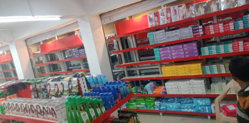 Retail Display Supermarket Rack Supplier And Rack Manufacturer in Andhra Pradesh, This is the interior of a cosmetics or personal care store. The store has bright fluorescent lighting on the ceiling along with ceiling fans. The walls are painted white, and there are prominent red-colored shelving units and display racks throughout the space. The shelves are well-organized and stocked with various personal care products. There are multiple levels of shelving, with the higher shelves containing boxed items in different colors - purple, teal, and yellow packaging can be seen. The lower display areas have bottles and containers, including shampoo or hair care products in blue, green, and other colored bottles. On the front shelves, there are hair dye or coloring products with pictures of models displaying different hairstyles. The store appears to be clean and well-maintained, with a modern retail layout designed to showcase the products effectively.