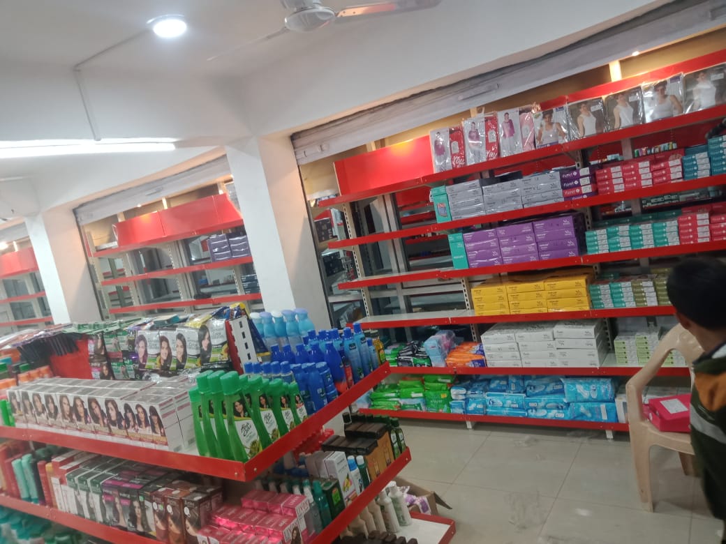 Retail Display Supermarket Rack Supplier And Rack Manufacturer in Andhra Pradesh, This is the interior of a cosmetics or personal care store. The store has bright fluorescent lighting on the ceiling along with ceiling fans. The walls are painted white, and there are prominent red-colored shelving units and display racks throughout the space. The shelves are well-organized and stocked with various personal care products. There are multiple levels of shelving, with the higher shelves containing boxed items in different colors - purple, teal, and yellow packaging can be seen. The lower display areas have bottles and containers, including shampoo or hair care products in blue, green, and other colored bottles. On the front shelves, there are hair dye or coloring products with pictures of models displaying different hairstyles. The store appears to be clean and well-maintained, with a modern retail layout designed to showcase the products effectively.